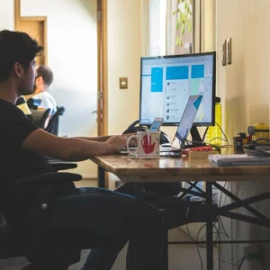 Open space bureau avec personne à lordinateur, écran, laptop, smartphone et accessoires de bureau.
