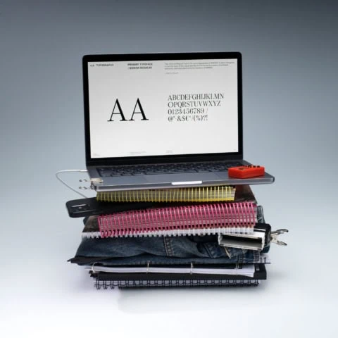 Ordinateur portable sur pile de cahiers et jeans, écran typographie, bureau improvisé.