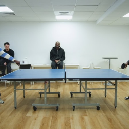 Match de ping-pong entre hommes en salle de loisirs, table, raquettes et spectateurs.
