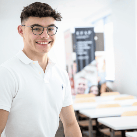 Portrait détudiant souriant en polo blanc avec lunettes dans une salle de classe lumineuse.