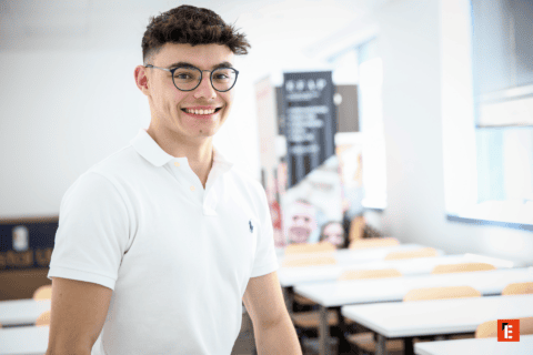 Portrait détudiant souriant en polo blanc avec lunettes dans une salle de classe lumineuse.
