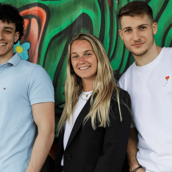 Portrait de groupe : trois jeunes souriants devant un graffiti coloré.