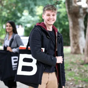 Portrait d’homme souriant avec sac noir «B» en toile, parc arboré automne.