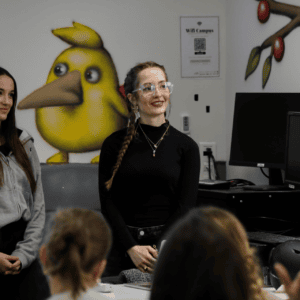 Jeunes enseignantes présentant devant étudiants, salle avec mur peint oiseau jaune et ordinateurs.