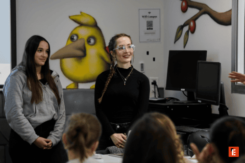 Jeunes enseignantes présentant devant étudiants, salle avec mur peint oiseau jaune et ordinateurs.