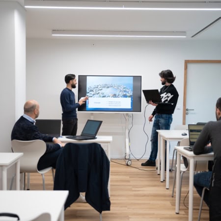 Présentation en petite salle de réunion avec écran, formateurs et participants sur ordinateurs portables.