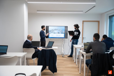Présentation en petite salle de réunion avec écran, formateurs et participants sur ordinateurs portables.