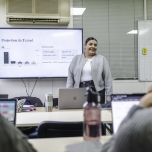 Présentatrice en salle de réunion devant écran, MacBook et participants concentrés.