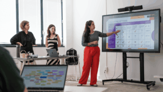 Réunion déquipe : trois femmes présentant un tableau dévaluation sur grand écran.