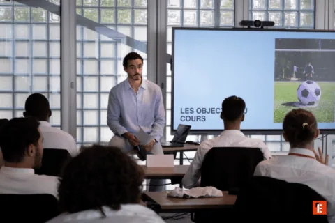 Réunion de formation en salle avec présentateur, écran, participants, coaching sportif et visioconférence.
