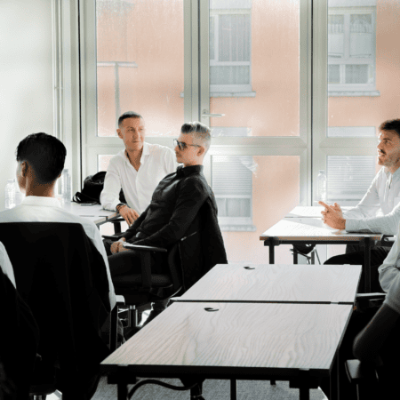 Réunion professionnelle en salle de classe lumineuse avec hommes, bureaux en bois et grandes fenêtres.