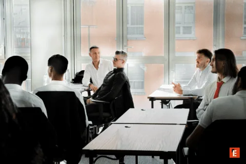 Réunion professionnelle en salle de classe lumineuse avec hommes, bureaux en bois et grandes fenêtres.