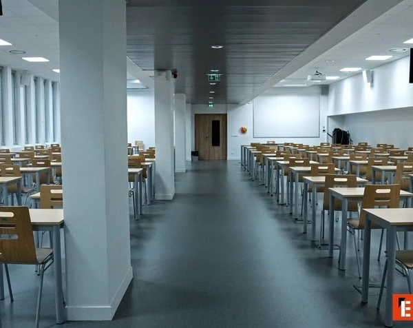 Salle dexamen vide avec rangées de bureaux et chaises en bois, fenêtres et lumière naturelle.