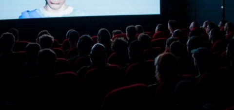 Spectateurs dans une salle de cinéma aux sièges rouges, écran lumineux.