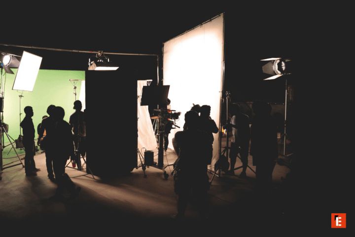 Studio de tournage avec écran vert, grande diffusion lumineuse, équipe technique et caméra en silhouette.