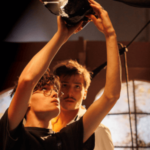 Technicien éclairage ajustant un projecteur sur plateau, assistant en arrière-plan, ambiance chaleureuse.