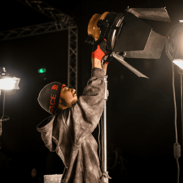 Technicien éclairage studio réglant un projecteur sur plateau de tournage sombre.