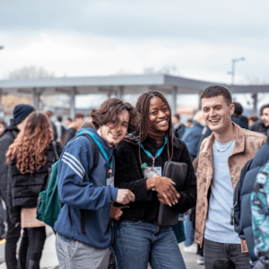 Trois jeunes souriants au arrêt de transport, foule hivernale, manteaux, badges et abri moderne.