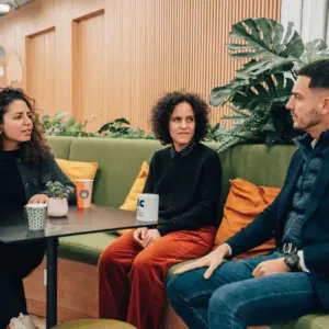 Trois personnes en réunion informelle sur canapé vert, plantes, café et ambiance collaborative.