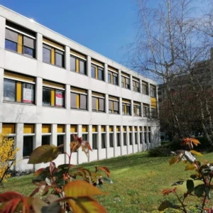 Bâtiment moderne trois étages façade blanche, fenêtres jaunes, pelouse et arbres.