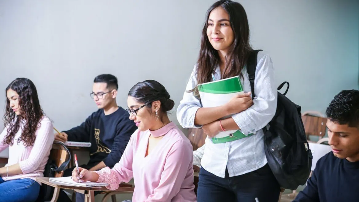 Élève debout tenant un carnet dans une classe studieuse avec élèves assis.