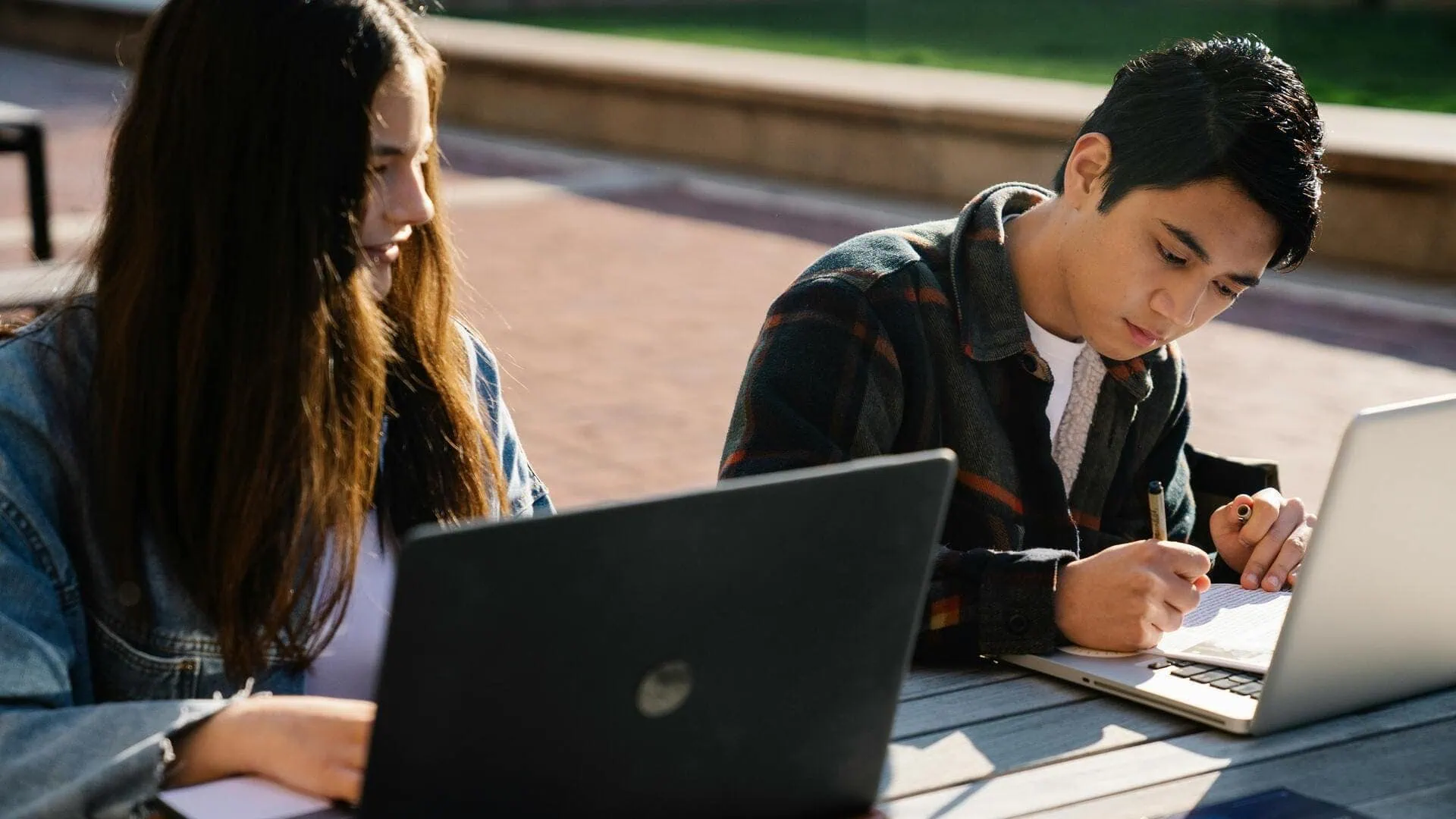 Étudiants travaillant dehors avec ordinateurs portables et cahiers, prise de notes au soleil.