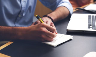 Homme écrivant une note au stylo près d’un ordinateur portable sur un bureau.