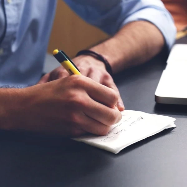 Homme écrivant une note au stylo près d’un ordinateur portable sur un bureau.
