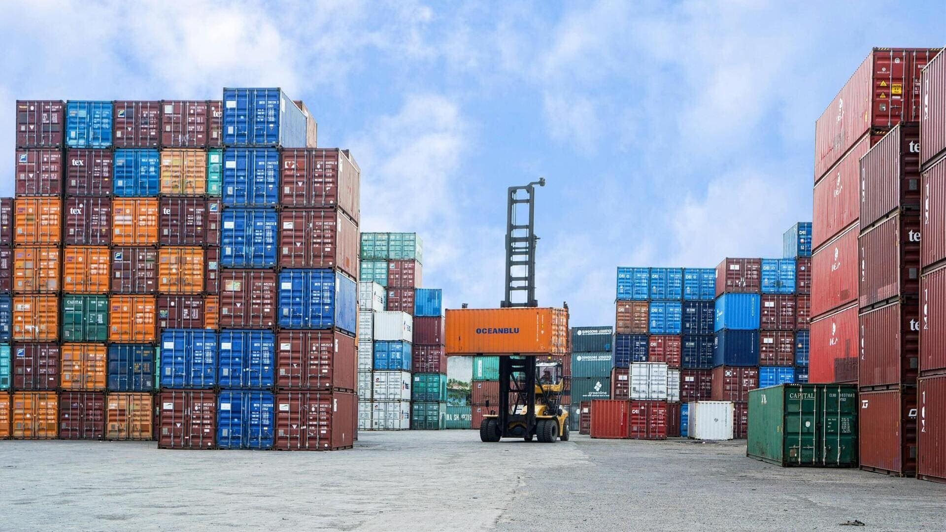 Parc de conteneurs portuaire, engin de manutention soulevant conteneur orange, piles empilées.