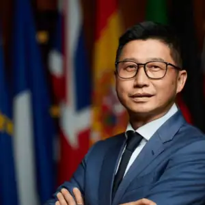 Portrait professionnel dun dirigeant en costume, lunettes et cravate, drapeaux flous en arrière-plan.