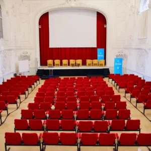 Auditorium universitaire vide avec scène, grand écran et rangées de sièges rouges.