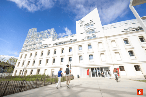 Bâtiment mixte pierre et verre, parvis et entrée animés par piétons et cycliste.