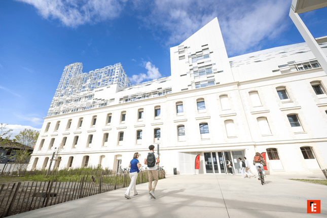 Bâtiment mixte pierre et verre, parvis et entrée animés par piétons et cycliste.