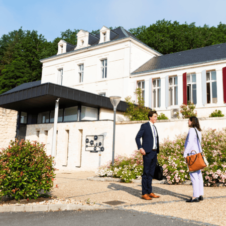 Bâtiment en pierre avec façade élégante, entrée moderne, jardins fleuris et professionnels en conversation.