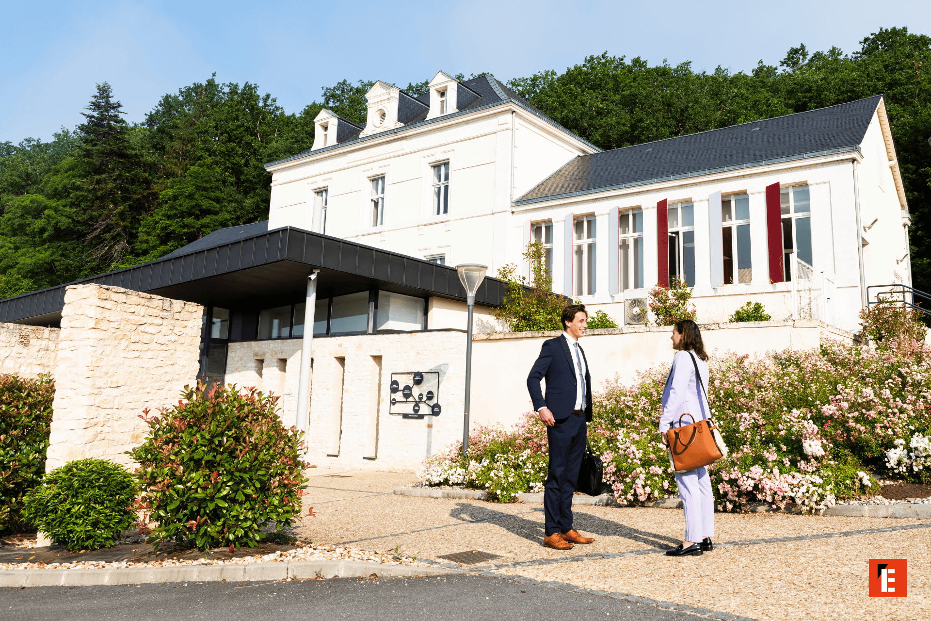 Bâtiment en pierre avec façade élégante, entrée moderne, jardins fleuris et professionnels en conversation.