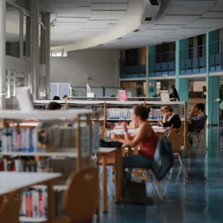 Bibliothèque moderne lumineuse avec grandes fenêtres, tables détude, étagères et étudiants concentrés.