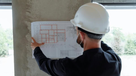 Ouvrier casqué sur chantier vérifiant plan architectural contre mur en béton.