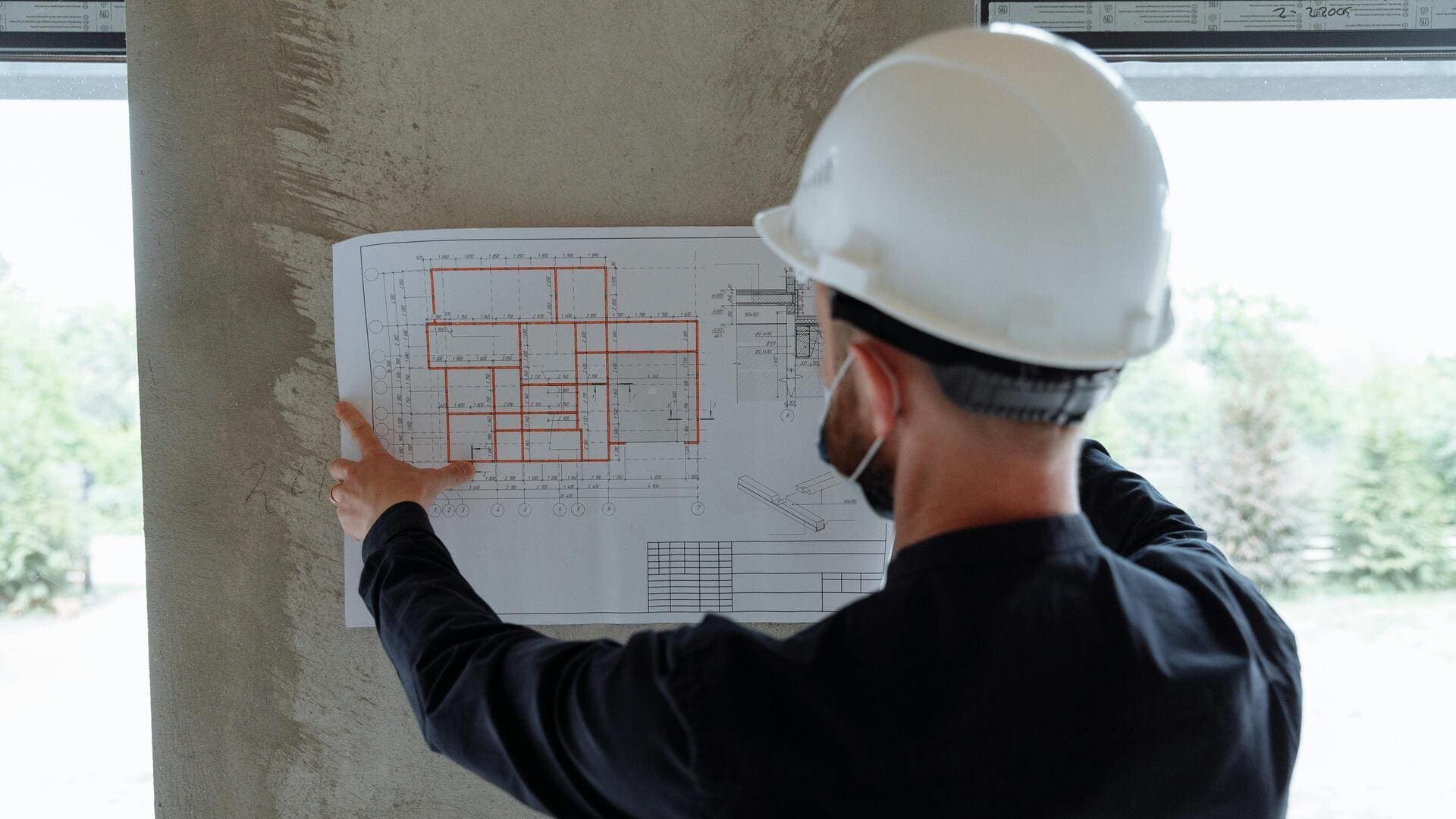 Ouvrier casqué sur chantier vérifiant plan architectural contre mur en béton.
