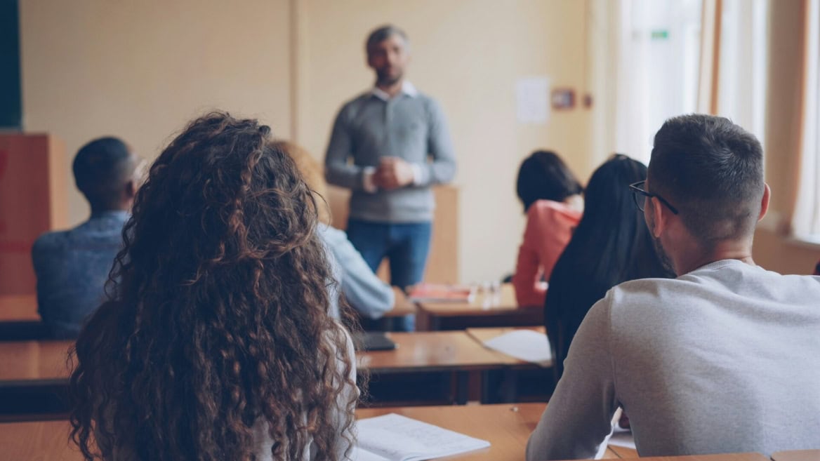 Classe avec élèves de dos, enseignant flou devant, prise de notes, lumière naturelle.