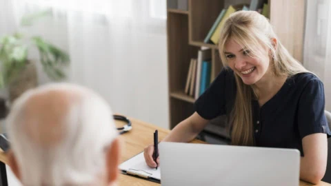 Infirmière souriante en consultation avec personne âgée, stéthoscope et ordinateur sur table.
