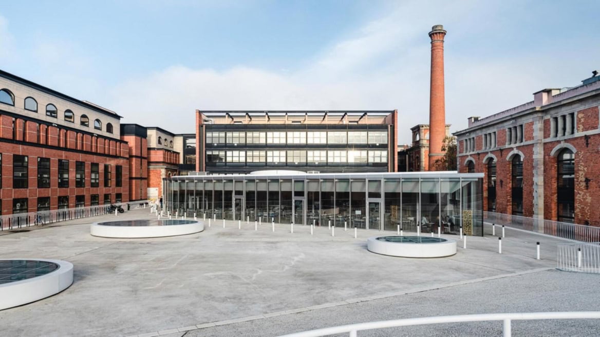 Cour urbaine rénovée avec pavillon en verre, café, bâtiments industriels et cheminée.