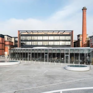 Cour urbaine rénovée avec pavillon en verre, café, bâtiments industriels et cheminée.