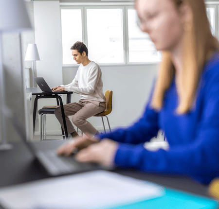 Coworking lumineux avec personne concentrée sur ordinateur portable, fauteuil jaune près des fenêtres.