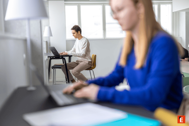 Coworking lumineux avec personne concentrée sur ordinateur portable, fauteuil jaune près des fenêtres.