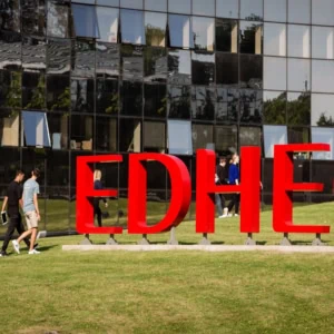 Signe rouge EDHEC sur pelouse de campus, étudiants en mouvement devant bâtiment vitré.