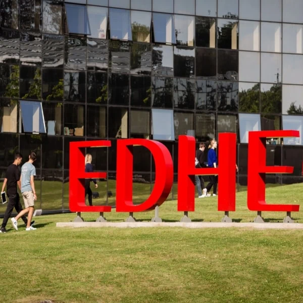 Signe rouge EDHEC sur pelouse de campus, étudiants en mouvement devant bâtiment vitré.