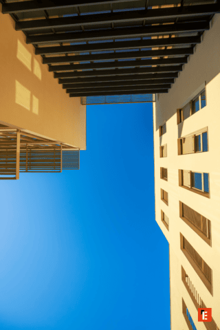 Architecture verticale entre bâtiments : façades sablées, lames métalliques, ciel azur et ombres géométriques.