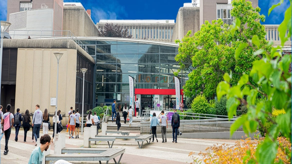 Entrée de campus : atrium vitré, étudiants, marches et végétation devant bâtiment universitaire.