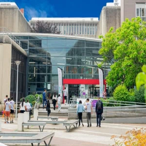 Entrée de campus : atrium vitré, étudiants, marches et végétation devant bâtiment universitaire.
