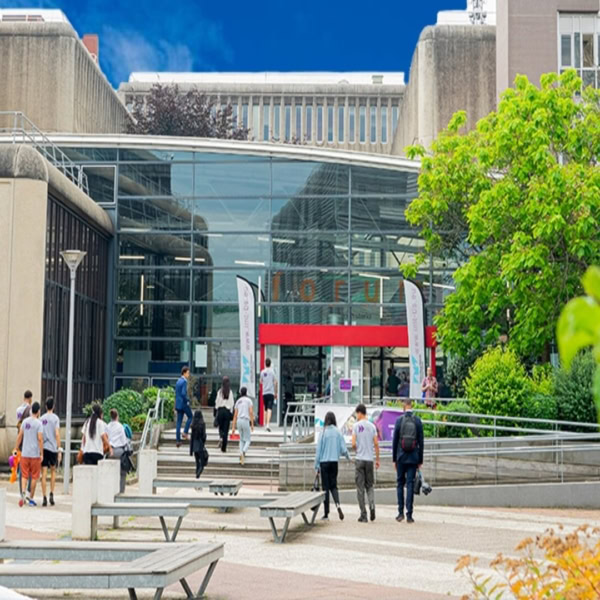 Entrée de campus : atrium vitré, étudiants, marches et végétation devant bâtiment universitaire.